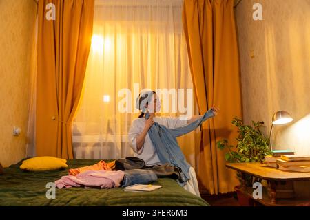 Kaukasische Frau faltet Wäsche in ihrem Zimmer, während die Sonne untergeht. Sie ordnet die Gegenstände sorgfältig an. Konzept der Raumorganisation. Niedriger Winkel, Weitwinkelaufnahme. Stockfoto