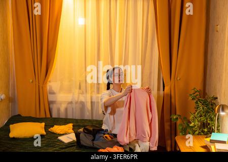 Kaukasische Frau faltet Wäsche in ihrem Schlafzimmer bei warmem Sonnenlicht am Abend. Sie organisiert ihren persönlichen Raum. Konzept für ein sauberes Leben. Mittelformat. Stockfoto