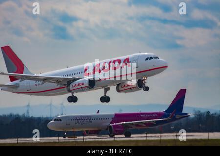 Eine Lauda Airbus A320 steigt nach dem Start direkt über einer am Boden rollenden Wizz Air Airbus A320 auf, wobei Windturbinen am Horizont im Hintergrund zu sehen sind Stockfoto