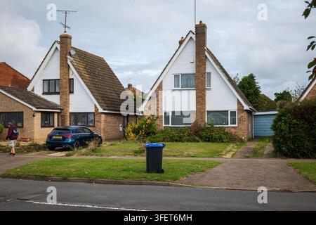 Wohnstraße in Northampton mit zwei freistehenden A-Förmigen Häusern mit Rasen, Einfahrtenparkplatz, einem blauen Auto, Garage und einem Rollcontainer im vorderen Garten Stockfoto