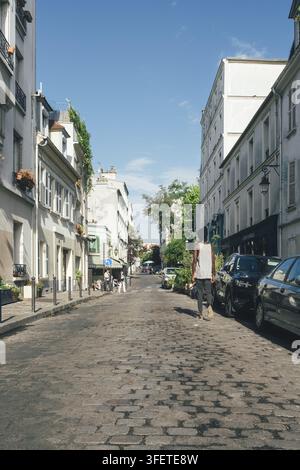 Blick auf eine charmante, gepflasterte Straße im historischen Viertel Montmartre mit charakteristischen alten Gebäuden, geparkten Autos und einer Person, die unter einem hellen Stockfoto
