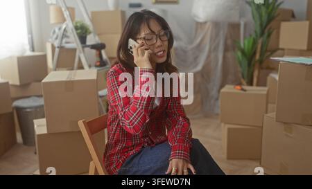 Frau, die auf einem Stuhl sitzt und Telefon an Ohr hält, zwischen Umzugskartons aus Pappe, Leiter und Topfpflanze im Gebäude; Erwartung des bewegenden Tages. Stockfoto