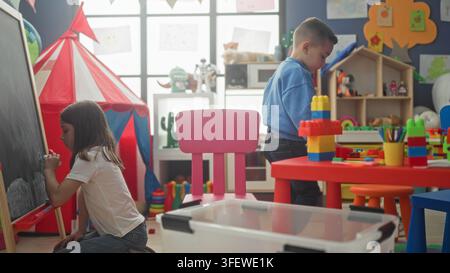 Junge baut einen Blockturm und Mädchen malt an einer Kreidetafel in einem Gebäude-Spielzimmer; Neugier-Kooperation. Stockfoto