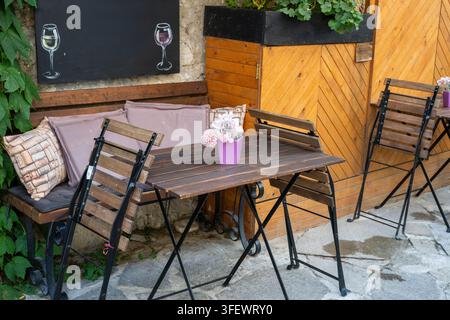 Eine gemütliche Außenterrasse eines Cafés mit einem Holztisch, Klappstühlen, einer Sitzbank mit Kissen und einem Kreidetafel-Schild, das Weinglaszeichnungen zeigt. Stockfoto