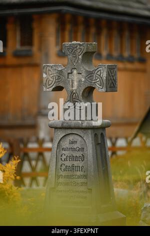 Ein verwitterter steinerner keltischer Kreuz-Grabstein steht auf einem Friedhof mit einer Holzkirche im Hintergrund. Stockfoto