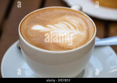 Eine Nahaufnahme von frisch gebrühtem heißem Kaffee mit zarter Farnblatt-Latte-Art in einer weißen Tasse auf einem passenden Unterteller. Stockfoto