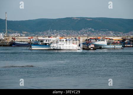 Eine Vielzahl von kleinen Fischerbooten und einem weißen Motorboot sind an einem langen Betonpier in einem ruhigen blauen Hafen festgemacht, mit einer Küstenstadt und sanften Hügeln Stockfoto