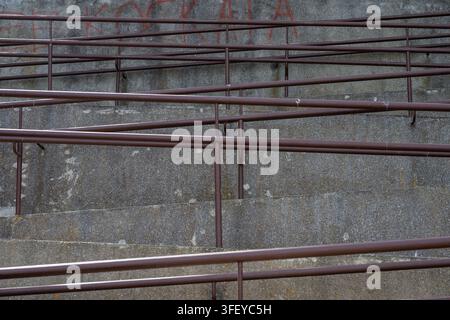Ein Netzwerk aus sich kreuzenden braunen Metallgeländern säumt eine im Zickzack verlaufende Beton-Zugangsrampe vor einer verwitterten grauen Wand. Stockfoto