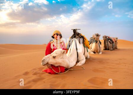 Lächelnde junge lateinamerikanische Frau in rotem marokkanischem Kleid und goldenem Kopftuch steht neben einem weißen Kamel in goldenen Wüstendünen von Marrakech. Der warme Sonnenuntergang weich Stockfoto