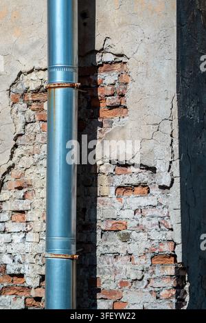 Metallabflussrohr an einer alten rissigen Gipswand mit freiliegendem rotem Backstein, Verwitterung und Rost. Stockfoto