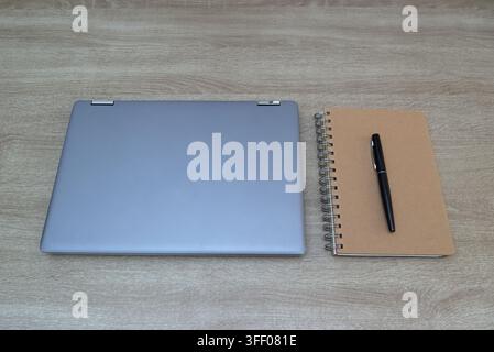 Sauberer Flat-Lay-Blick auf einen geschlossenen grauen Laptop neben einem Spiralnotizbuch und einem schwarzen Stift auf einem hellen Holzschreibtisch, der Organisation, Fokus und Stockfoto