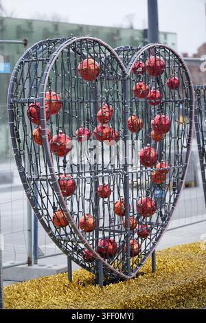 Eine große herzförmige Skulptur aus dunklem Metallmaschengewebe ist gefüllt mit glänzenden roten und orangefarbenen reflektierenden Zierkugeln und Strängen kleiner Lichter Stockfoto