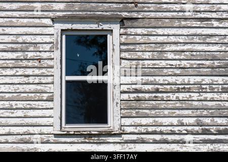Ein Wohnfenster befindet sich links von einem breiten Kopierraum an einer gealterten Holzschindelnwand mit abblätternder weißer Farbe und freiliegendem verwittertem Holz. Stockfoto