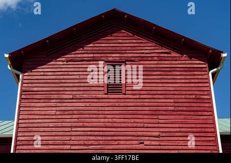 Symmetrische Ansicht des Giebels eines alten roten Holzhauses mit verwittertem horizontalem Seitenverkleidungs und einem kleinen Lüftungsschacht unter einem tiefblauen Himmel in Stockfoto