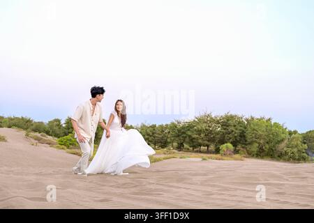 Ein fröhliches indonesisches südostasiatisches Paar geht Hand in Hand über sandige Dünen. Die Frau trägt ein fließendes weißes Hochzeitskleid und der Mann ist in einem lässigen Anzug Stockfoto