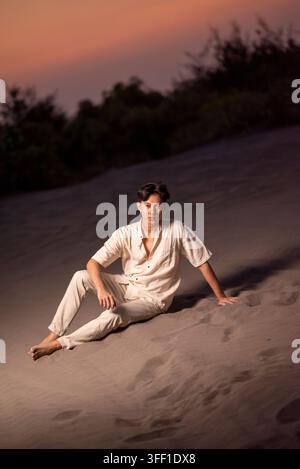 Ein junger indonesischer Südostasiate in einem stilvollen weißen Leinenhemd und einer Leinenhose sitzt entspannt auf einer Sanddüne und posiert unter einem dramatischen Abendhimmel bei der Dämmerung Stockfoto