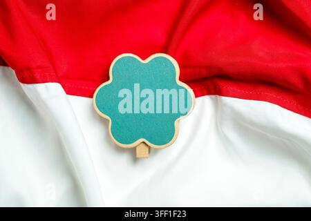 Ein kleiner leerer grüner, blumenförmiger Holztafeln-Clip, der auf einem welligen roten und weißen indonesischen Nationalflaggenstoff platziert ist, geeignet für Kopierplatz Stockfoto