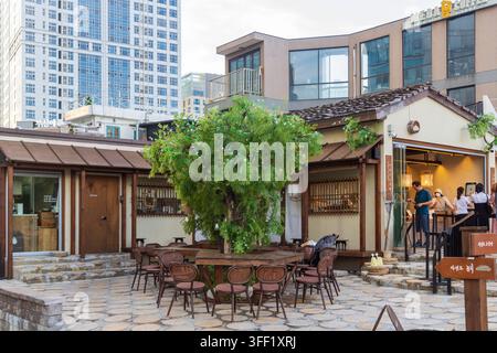 Eine Außenterrasse eines Cafés mit Holztischen, Stühlen, Bäumen und Gästen steht unter einem modernen Hochhaus mit Glasfassade. Stockfoto