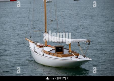 Nahaufnahme eines kleinen weißen Segelboots, das auf ruhigem Wasser schwebt. Klassisches Segelboot mit Holzdetails, erhobenem Mast und eingerollter Segelbahn, fotografiert von Stockfoto