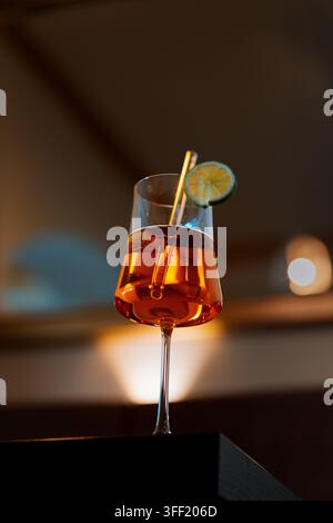 Aperol-Spritz-Cocktail in einem Weinglas mit Orangenscheibe und Strohhalm, warmes Abendlicht der Bar und dunkler Bokeh-Hintergrund Stockfoto