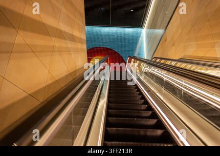 Blick nach oben zu einer beleuchteten Rolltreppe in einer modernen Station, die zu einer farbenfrohen Kachelwand mit rotem und blauem kreisförmigem Kunstwerk führt Stockfoto