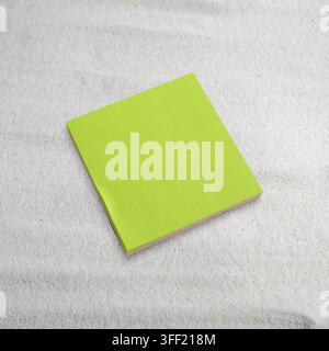 Ein quadratischer limettengrüner Haftnotizblock, der auf feinem weißem Sand ruht und ein minimalistisches Konzept bildet. Platz für Text oder Beispiel Stockfoto