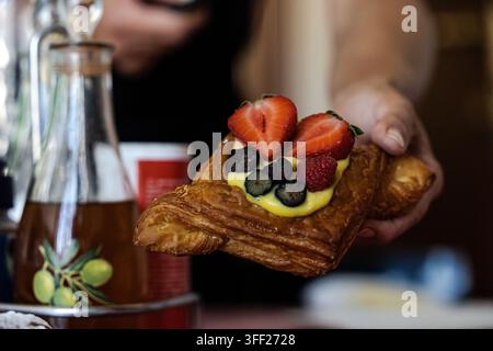 Eine Hand hält eine frische, blättrige Blätterteigtarte, die mit reichhaltiger Vanillesauce, geschnittenen Erdbeeren, Blaubeeren und einer frischen Himbeere garniert ist. Stockfoto