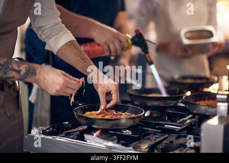 Nahaufnahme der Hand eines Kochs, der Speck in Eier gibt, während er Frühstück zubereitet. Koch in einer professionellen modernen Küche, Restaurantkonzept Stockfoto