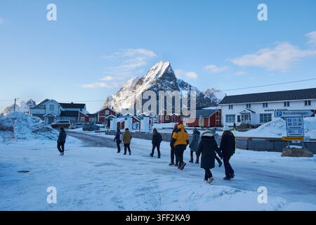 Schnee bedeckt eine ruhige Dorfstraße. Menschen gehen zusammen, gegen die Kälte eingepackt. Ein hoch aufragender, schneebedeckter Berg erhebt sich hinter den Häusern. Klares Blau s Stockfoto