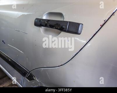 Nahaufnahme einer verwitterten silbernen Autotür mit erheblichen Schäden. Zeigt einen abgenutzten schwarzen Türgriff, Kratzer und abgeplatzte Farbe, betont das Alter und den Zustand des Fahrzeugs. Stockfoto