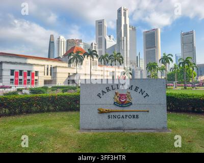 Stein mit der Aufschrift „Parliament Singapore“ neben dem nationalen Wappen und einem zeremoniellen Zepter. Parlamentsgebäude und Skyline im Hintergrund Stockfoto