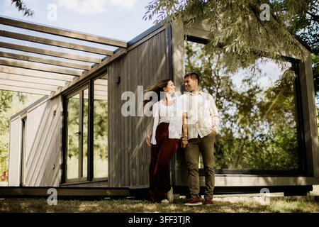 Ein glückliches Paar geht Hand in Hand, lächelt sich an, neben einem zeitgenössischen Haus. Stockfoto