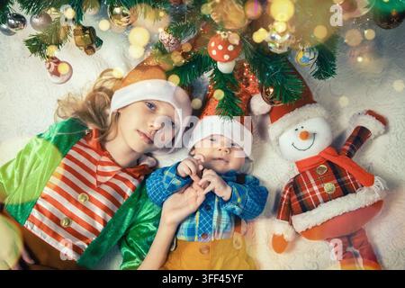 Zwei Kinder liegen unter einem Weihnachtsbaum, der mit bunten Ornamenten geschmückt ist und von Feiertagsdekorationen umgeben ist, während sie einen freudigen Moment teilen. Stockfoto