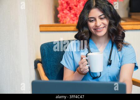 Zufriedene Ärztin genießt eine Kaffeepause, während sie am Laptop arbeitet Stockfoto