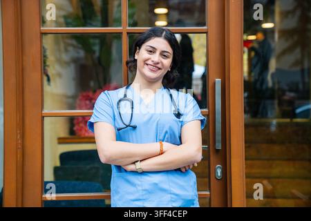 Porträt einer selbstbewussten Ärztin mit verschränkten Armen, die blaue OP-Kleidung und ein Stethoskop trägt. Ideal für Gesundheitsfachkräfte, medizinische Berufe, ho Stockfoto
