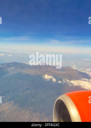 Atemberaubender Luftbildblick auf einen majestätischen Vulkan mit einem Kratersee, aufgenommen vom Flugzeugfenster aus, das den orangefarbenen Triebwerksbug zeigt. Stockfoto