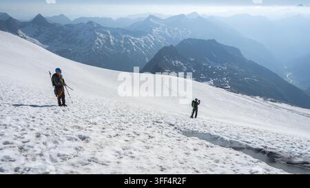 Zwei Bergsteiger steigen einen schneebedeckten Gletscher auf dem Grossvenediger in Österreich hinauf, mit dramatischem Alpenpanorama und nebligen Talblicken unten Stockfoto