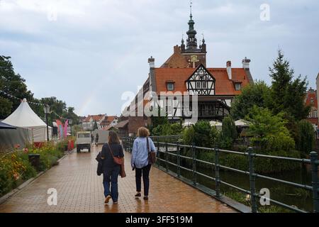 Menschen gehen über die Radunia-Kanalbrücke in Richtung des historischen Fachwerkgebäudes Dwór Młynarzy mit dem Turm der St.-Katharinen-Kirche im Hintergrund. Stockfoto