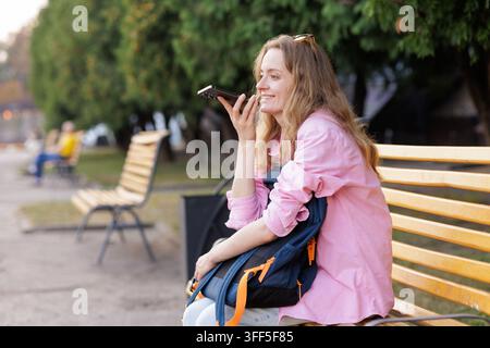 Junge Frau mit Rucksack sitzt auf einer Parkbank und nutzt den Sprachassistenten ihres Smartphones, was Reiseplanung, mobile Suche und alltägliche Outdoor-Kommunikation repräsentiert Stockfoto