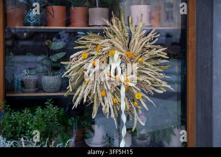 Dekorativer Kranz aus getrockneten Blumen, Weizenähren und Gras, der am Glasfenster eines Blumenladens hängt. Stockfoto