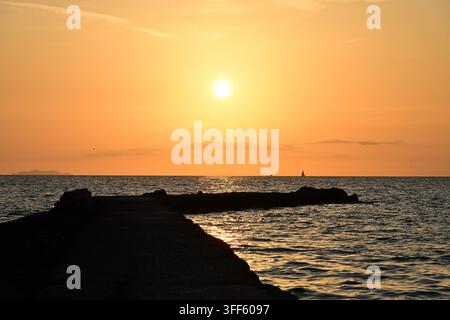 Warme goldene Sonne über ruhigem Ozean, Silhouette eines felsigen Piers, entferntes Segelboot am Horizont Stockfoto