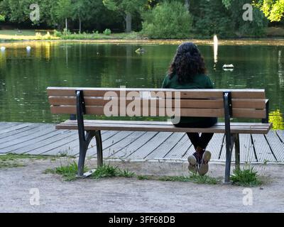 Eine einsame Person sitzt auf einer Holzbank und blickt über einen ruhigen Parksee mit einem fernen Brunnen. Fängt Themen der Einsamkeit, psychischen Gesundheit, Stockfoto