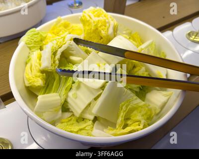 Eine Nahaufnahme einer weißen Keramikschale gefüllt mit frisch geschnittenem Pekinkohl, begleitet von Metallzangen an einem Buffet. Stockfoto