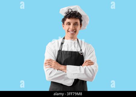 Gastronomieberuf und Hotellerie-Karriere, lächelnder junger Koch in weißer Uniform und schwarzer Schürze, der mit verschränkten Armen auf blauem Hintergrund steht Stockfoto
