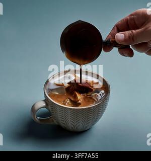 Heißes Espresso aus einem Metallkännchen in eine strukturierte Tasse mit Milch und Eis auf einem schieferblauen Hintergrund gießen Stockfoto