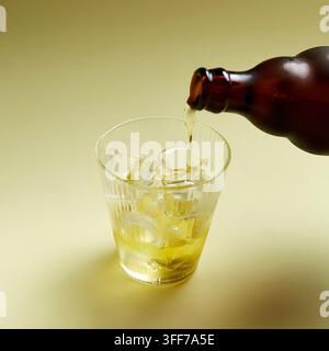 Goldenes Getränk aus einer braunen Flasche in ein geripptes Glas mit Eis auf einem hellgelben Hintergrund gießen Stockfoto