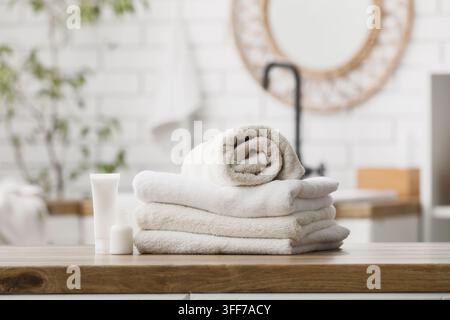 Wellness und Spa-Selbstpflege, Stapel sauberer weißer Handtücher und Hautpflegeprodukte auf einem hölzernen Badezimmertisch mit unscharfem Innenraumhintergrund Stockfoto
