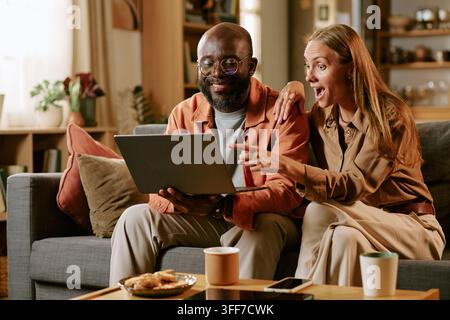 Verschiedenes Paar, afrikanisch-amerikanischer Mann und kaukasische Frau, die während der Black-Friday-Sale mit begeisterter Emotion online auf einem Laptop einkaufen. Visuelles Konzept für e Stockfoto
