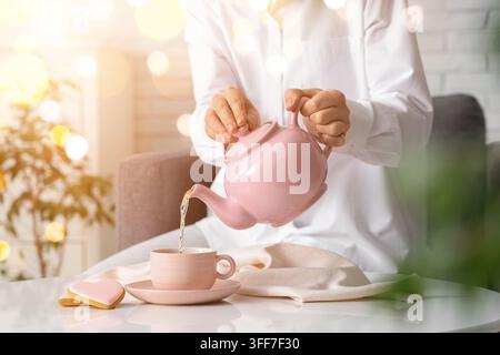 Selbstfürsorge und Achtsamkeit, Frau gießt Kräutertee in eine Tasse mit herzförmigen Keksen zur Entspannung und für die psychische Gesundheit zu Hause in einer gemütlichen Atmosphäre Stockfoto