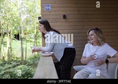 Schüssel mit frischen Kirschen auf einem Holzgeländer, zwei Frauen mit Getränken auf einem Sommerbalkon. Saisonobst und ein langsamer Nachmittag in einem Landhaus im f Stockfoto